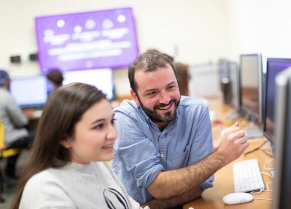 Jonah Warren sits at a table helping a female student with a video game project on a computer