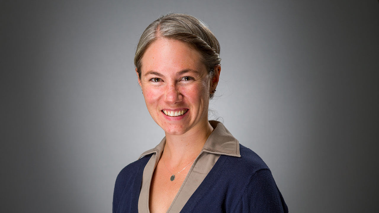 Kimbery DiGiovanni poses smiling for the camera, she is a white woman with blonde hair pulled back into a bun wearing a gray shirt and navy vest