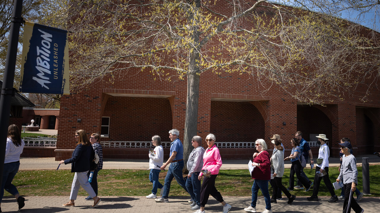 A group of people walk across campus under a few trees on a sunny day