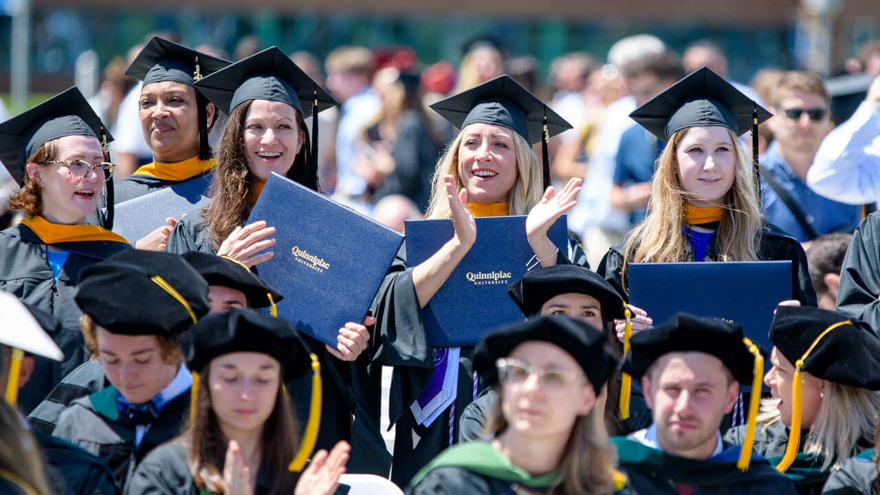 A group of graduates standing in the crowd at Commencement on the Quad, clapping for their fellow graduates.