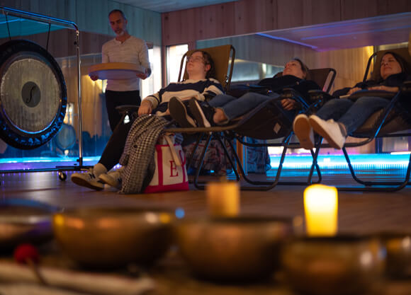 3 people relax in chairs in a dim room lit by candles while listening to someone play a gong