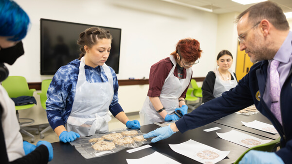 3 students in lab gear look at a specimen with a professor in a lab during academic exploration day